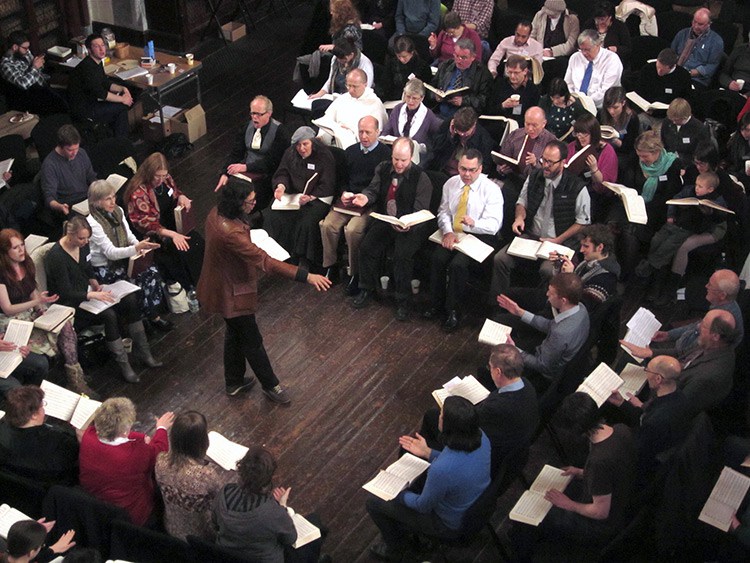 A photograph of a typical Sacred Harp singing: a singer stands on a dark wood floor as he leads a song from The Sacred Harp in the center of a hollow square featuring rows of seated singers with the songbook in their laps.